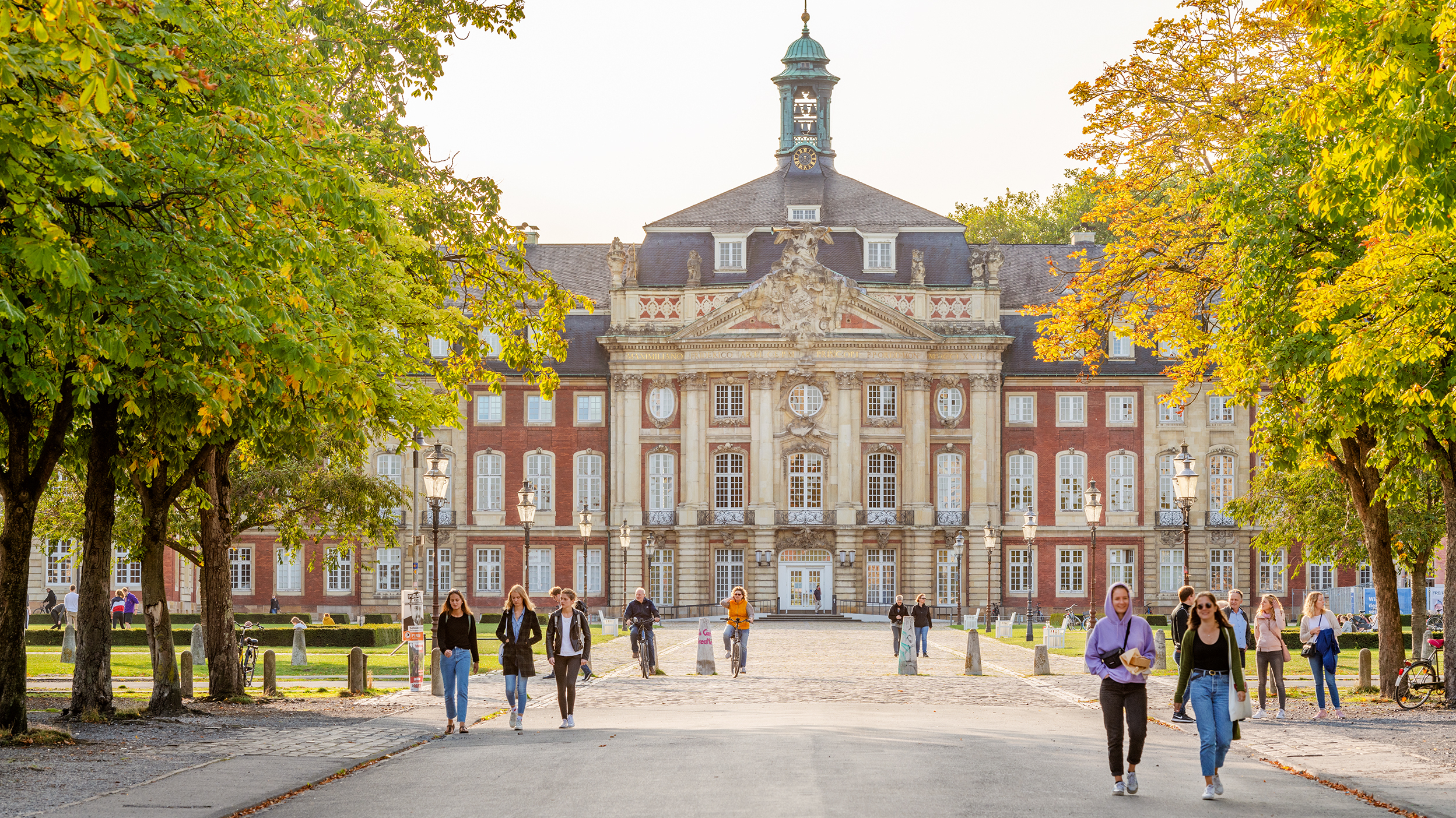 Das Schloss Münster in der Frontalansicht mit grünen Bäumen und Passanten im Vordergrund.
