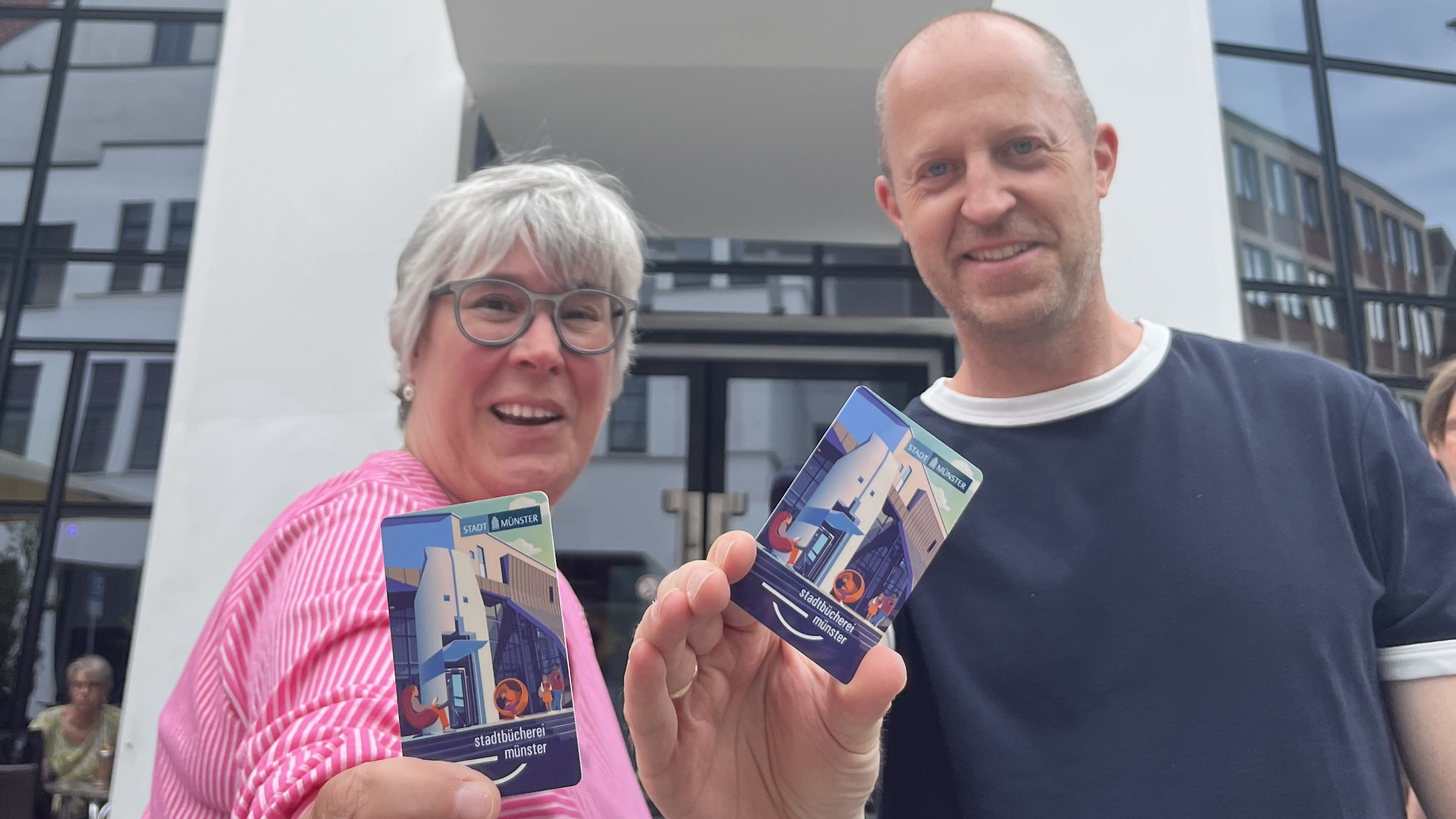 Cordula Gladrow, Leiterin der Stadtbücherei, und Grafiker Lars Wentrup halten den den neuen Büchereiausweis in die Kamera. Im Hintergrund ist die Bücherei zu sehen.