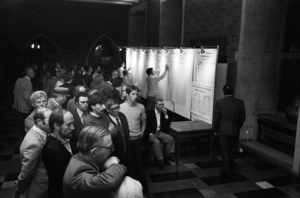 Mehrere Männer stehen in einem Raum dicht beieinander. Alle schauen auf die rechte Bildhälfte, wo vermutlich ein Fernseher aufgestellt war. Im Hintergrund notiert ein Mann Zahlen an einer Stellwand.