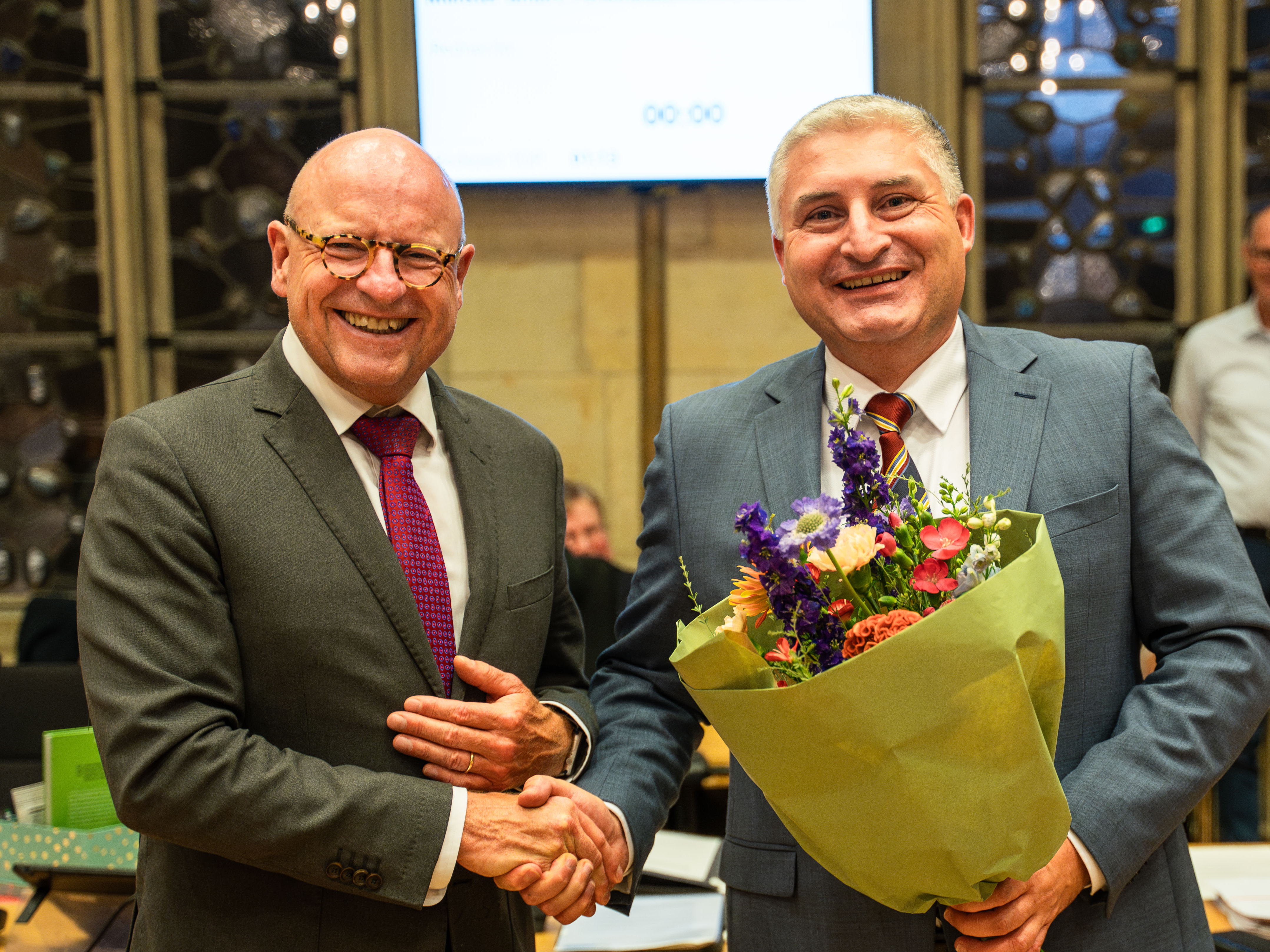 Zwei Männer schütteln sich die Hand; einer hält einen Blumenstrauß in der Hand