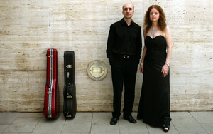 Die Gitarist*innen Ignacio Gea und Marisa Gómez stehen vor einer Mauer und blicken in die Kamera. Neben ihnen stehen zwei Gitarrenkoffer an die Wand gelehnt.