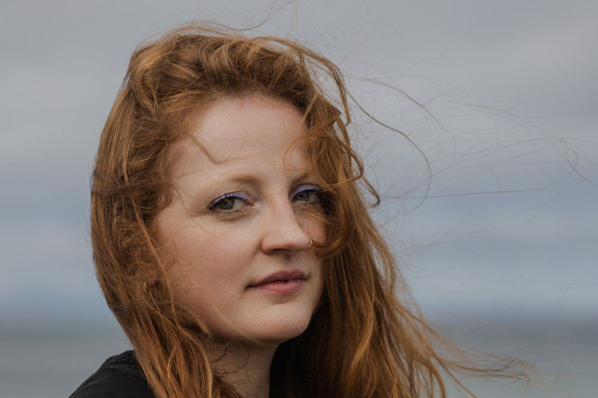 Frau mit langen roten Haaren sitzt am Meer und hält eine Gitarre