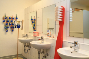 Blick in ein Kita- oder Schulbadezimmer mit drei Waschbecken und einem menschengroßen Modell einer roten Zahnbürste