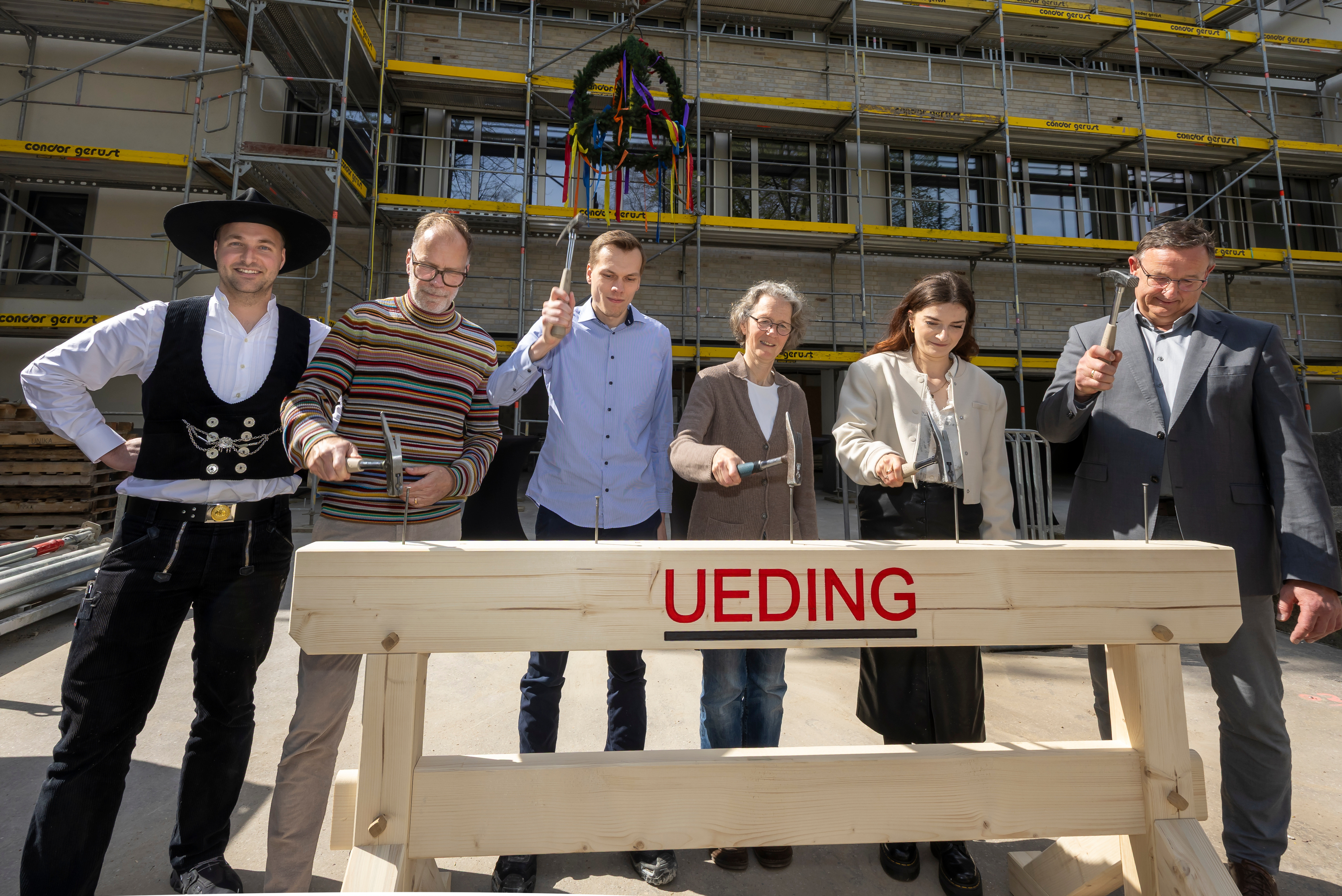 Sechs Personen schlagen jeweils mit einem Hammer auf einen Nagel in einem Balken.