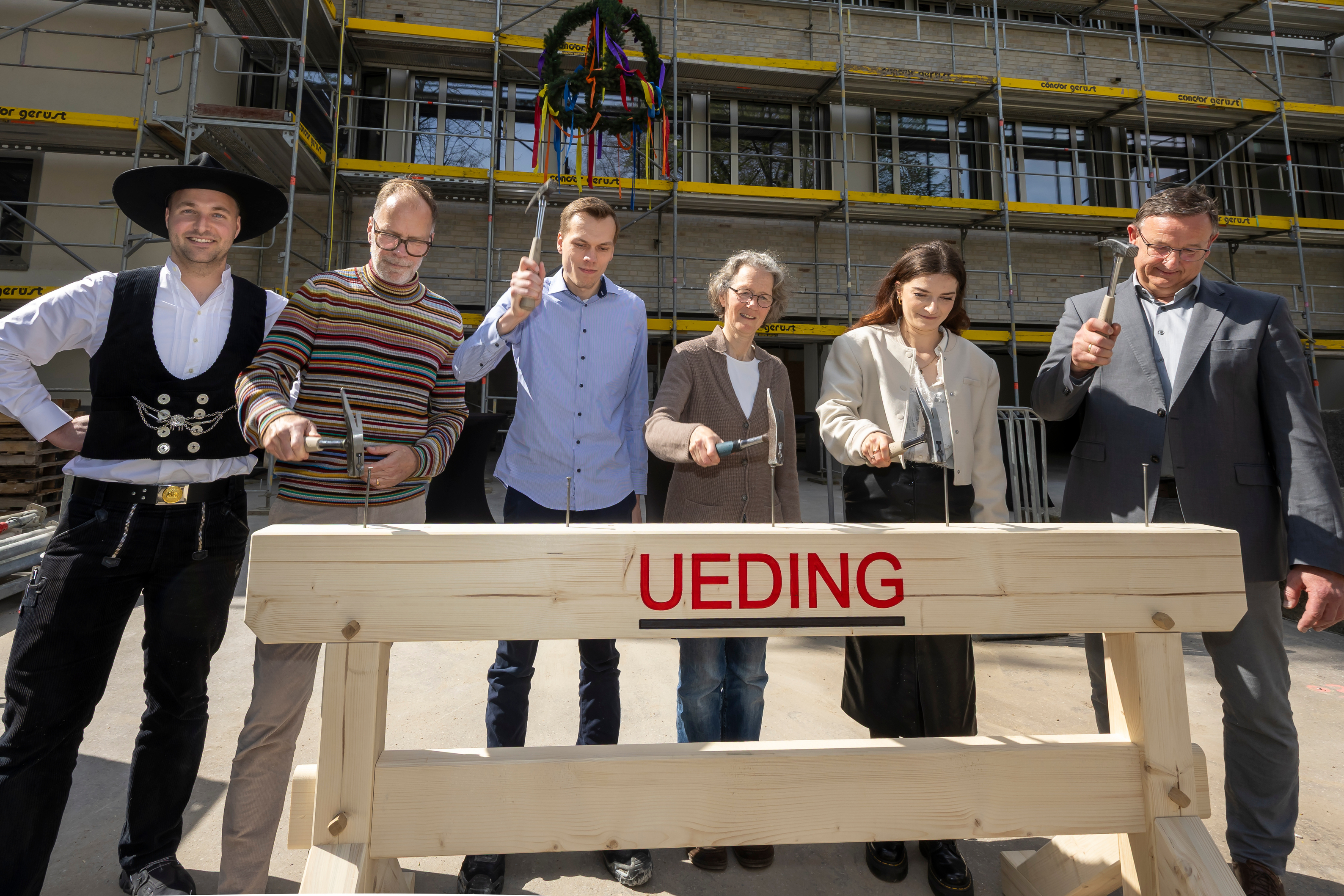 Sechs Personen schlagen jeweils mit einem Hammer auf einen Nagel in einem Balken.