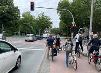 Viele Radfahrende stehen vor einer roten Ampel, ein Radfahrer hält sich an einem Ampeltrittbrett fest.