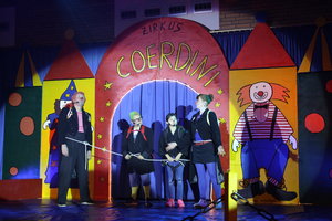 Ein Tor aus Pappe auf dem 'Zirkus Coerdini' zu lesen steht. Davor haben sich fünf Akteure in Kostümen nebeneinander aufgestellt.
