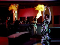 Eine belebte Bar mit zahlreichen Männern in Anzügen, einer tanzenden und einer leicht bekleideten Dame. Zwei Männer halten jeweils zwei brennende Flaschen in den Händen.