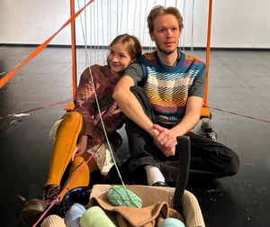 Eine Darstellerin und ein Darsteller sitzen auf dem Boden. Sie legt ihren Arm auf seine Schulter. Vor ihnen steht ein Korb mit Wolle.