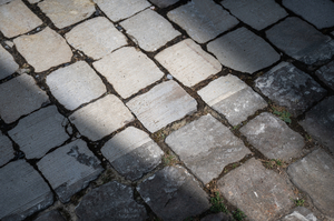 Das Foto zeigt Kopfsteinpflaster: Auf der einen Hälfte des Bildes ist es bereits geschliffen, d.h. es sind Rillenerkennbar und es ist heller und auf der anderen Hälfte des Bildes ist das Kopfsteinpflaster noch ungeschliffen