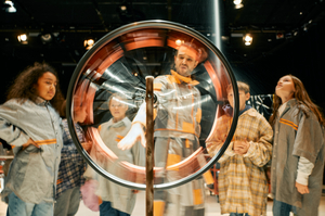 Ein Performer dreht ein Lotterierad. Hinter ihm stehen vier Jugendliche und schauen gespannt zu.