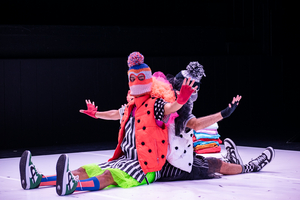 Zwei Performer sitzen mit ausgebreiteten Armen Rücken an Rücken auf dem Boden. Sie tragen bunte Kleidung und haben jeweils eine Pudelmütze auf dem Kopf.