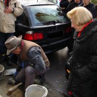 An einer Straße kniet ein Mann mit Handwerkszeug vor einem eingelassenen Stolperstein. Neben ihm steht eine Frau und blickt darauf, um sie herum stehen weitere Personen, die zusehen.