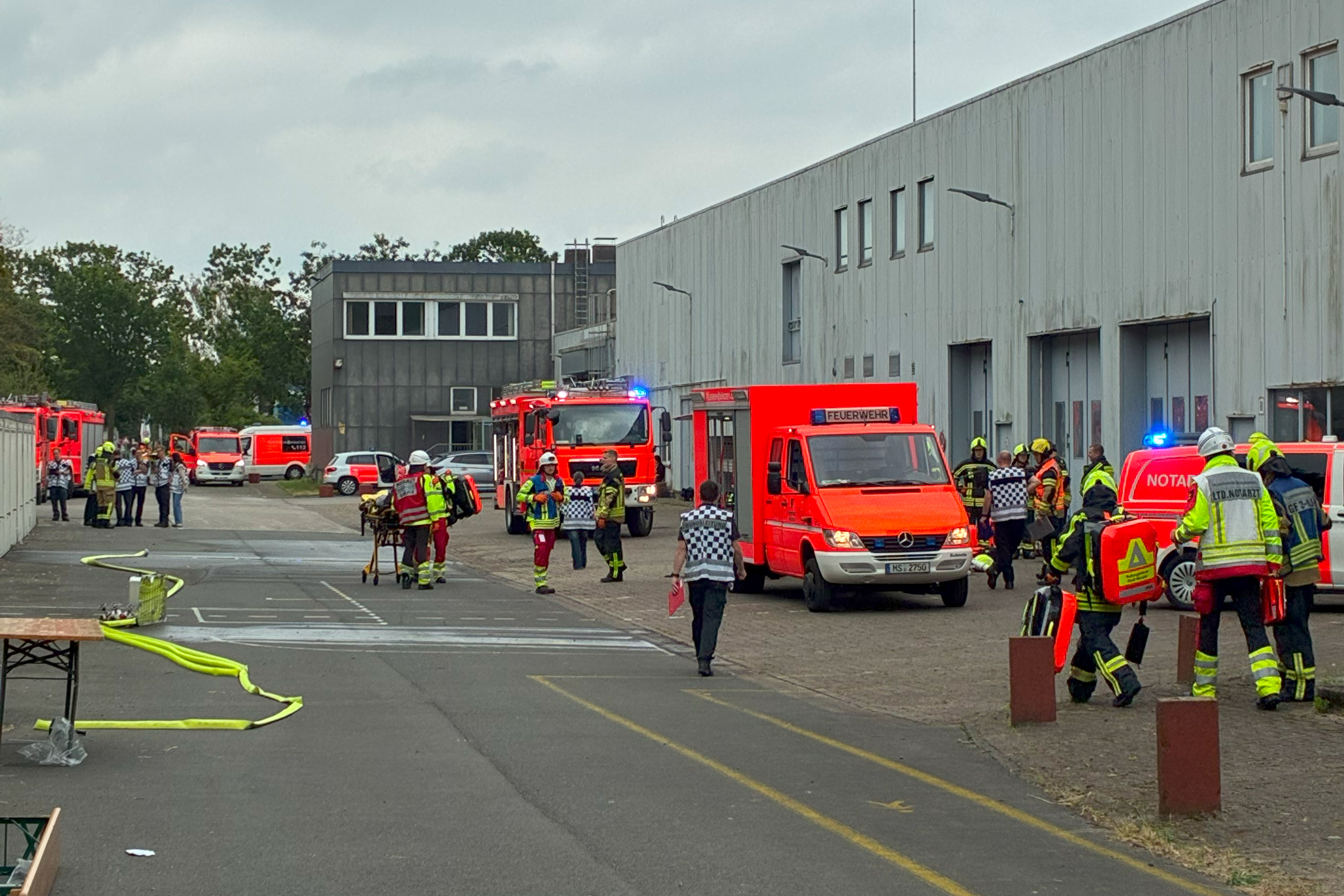 Eine große Halle auf einem weitläufigen Areal. Es befinden sich dort Mitarbeitende der Feuerwehr sowie Einsatzfahrzeuge und Equipment.