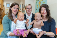 Drei Frauen halten zwei Babypuppen hoch, eine Puppe weist Merkmale einer Fetalen Alkoholspektrum-Störung auf.