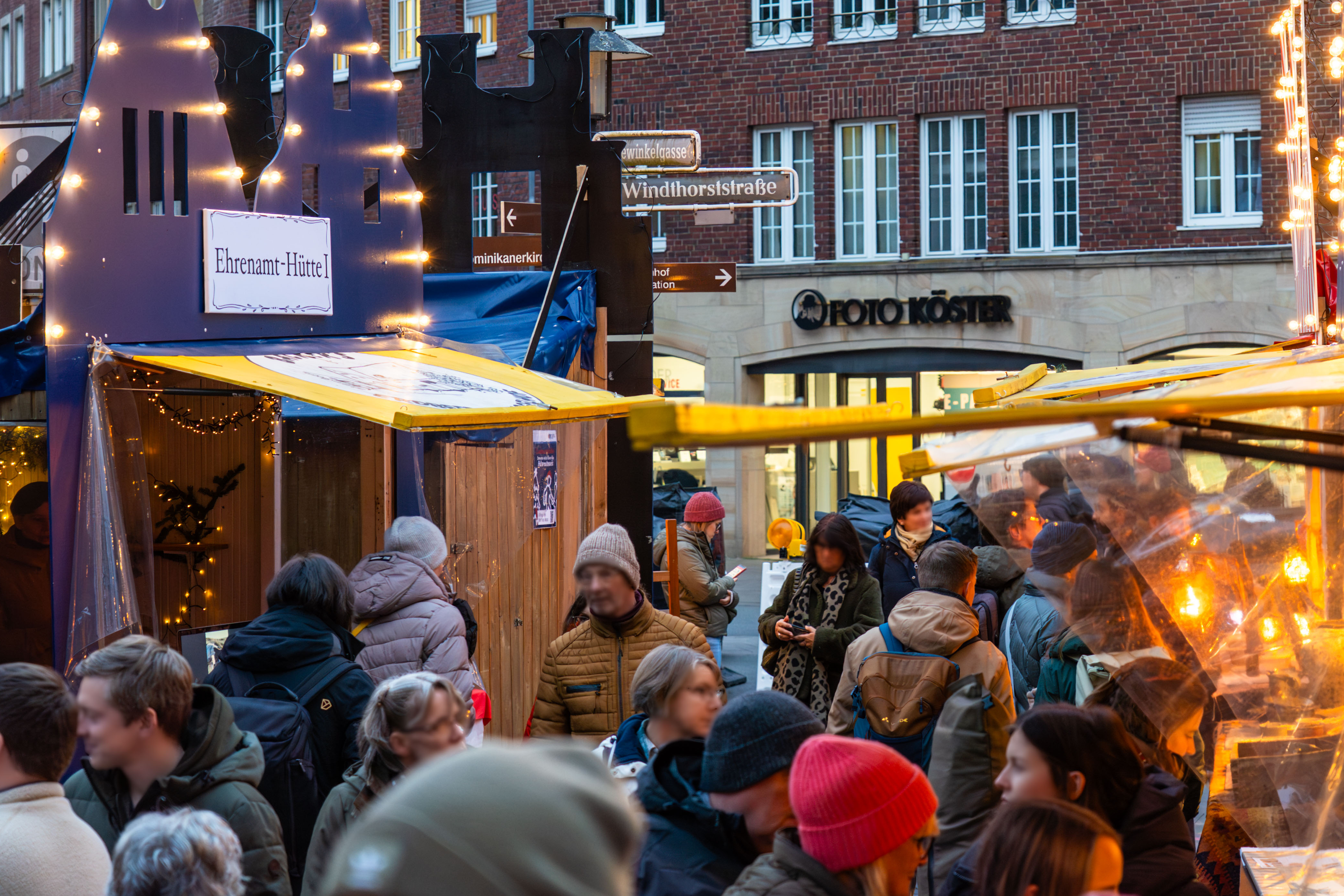 Der Weihnachtsmarkt am Harsewinkelplatz. Viele Menschen sind zwischen den Buden unterwegs, dazu gibt es festliche Beleuchtung.