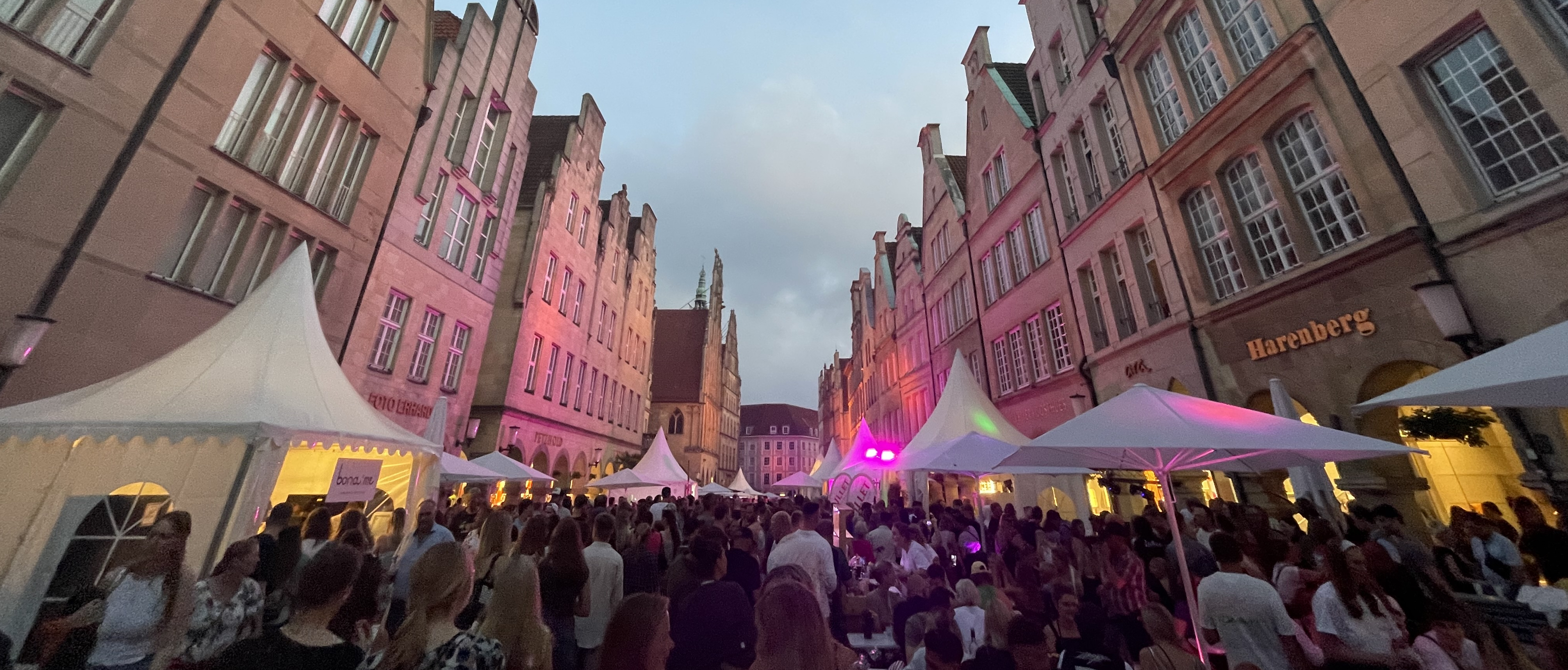 Blick in die Straße Prinzipalmarkt, links und rechts die Häuserfassaden, unten im Vordergrund eine feiernde Menschenmenge und weiße Pavillons am Straßenrand. Oben ein bewölkter Himmel.