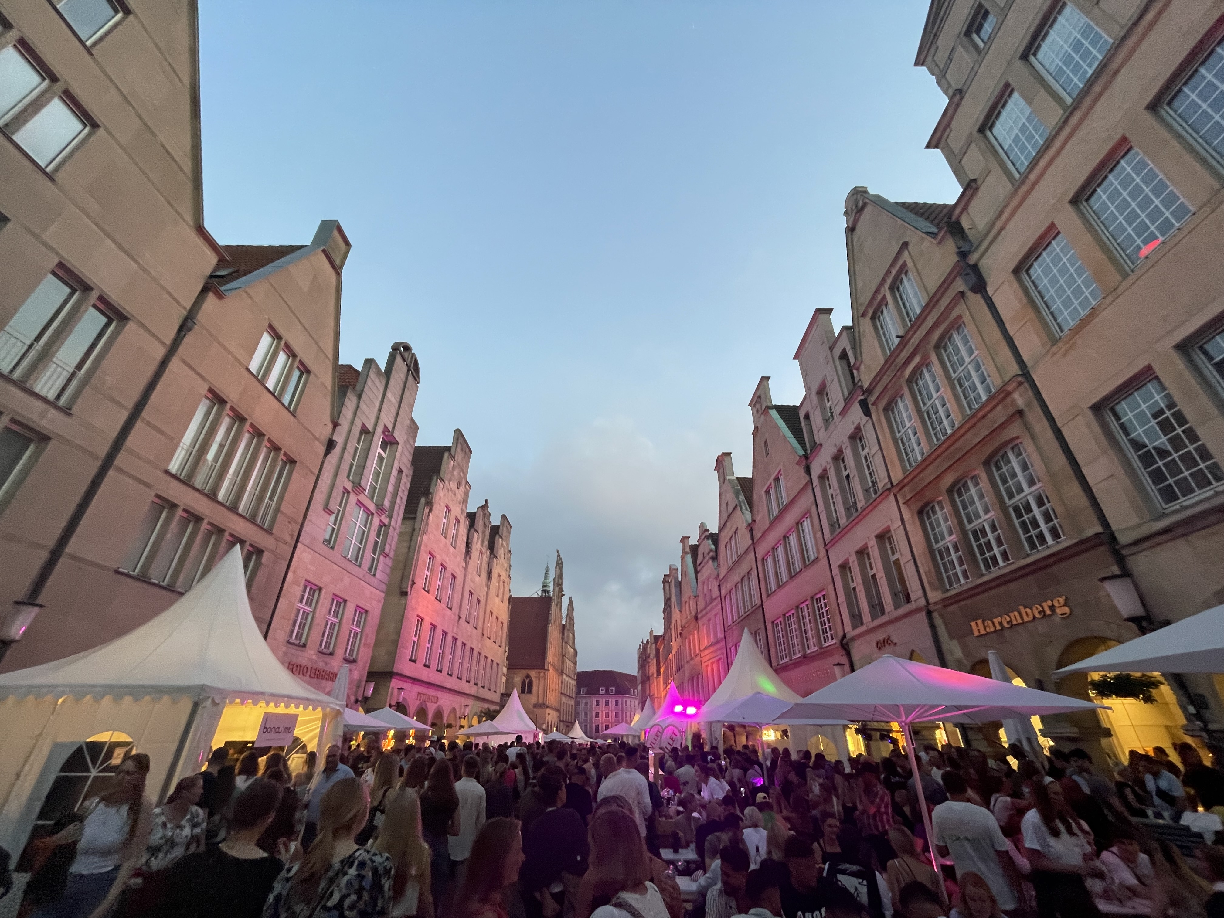 Blick in die Straße Prinzipalmarkt, links und rechts die Häuserfassaden, unten im Vordergrund eine feiernde Menschenmenge und weiße Pavillons am Straßenrand. Oben ein bewölkter Himmel.