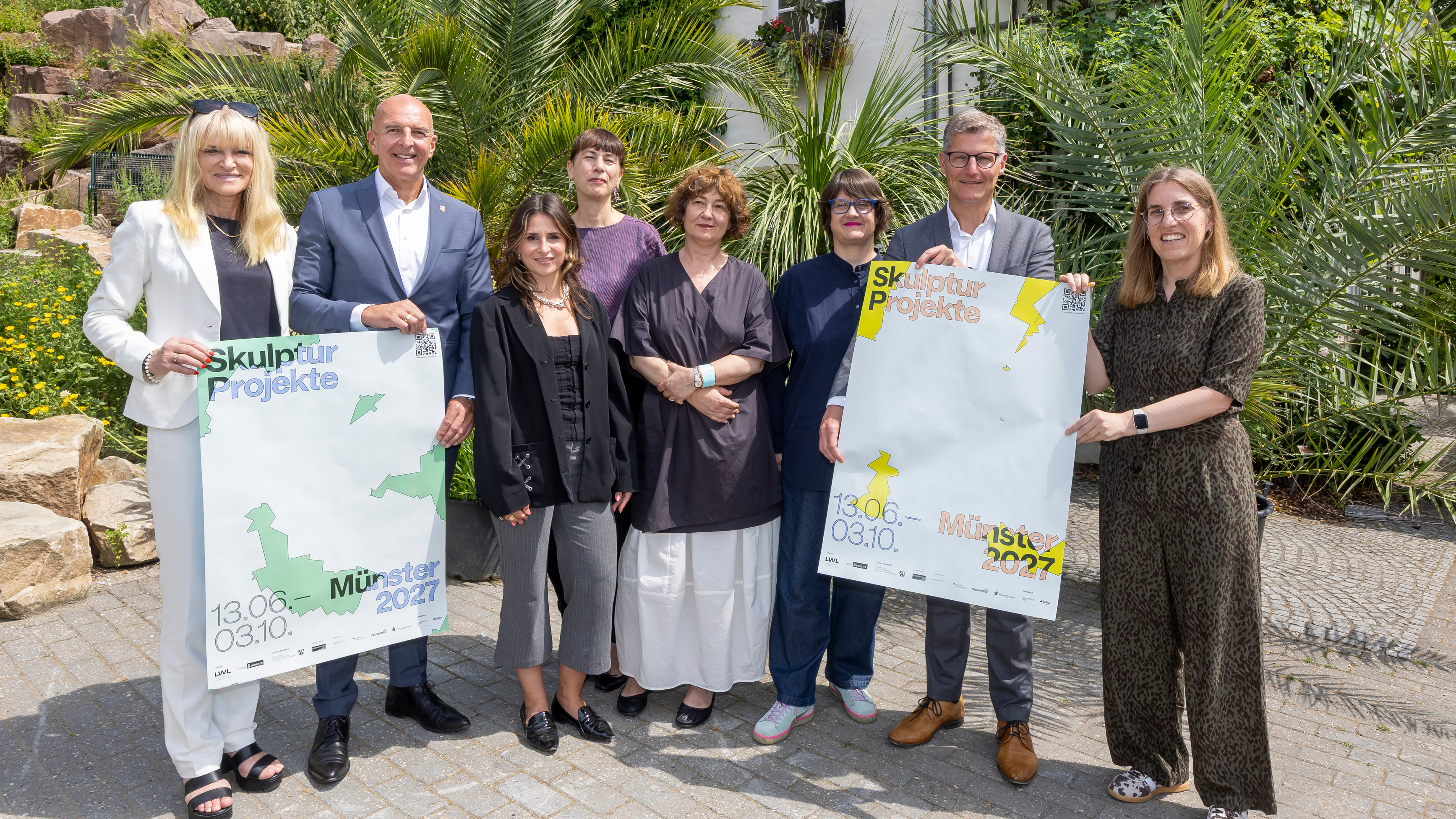 LWL-Kulturdezernentin Dr. Barbara Rüschoff-Parzinger, LWL-Landesdirektor Dr. Georg Lunemann, Verena Putzer (Projektleiterin Skulptur Projekte 2027), Ivet Ćurlin, Nataša Ilić und Sabina Sabolović (Kuratorinnenkollektiv WHW), Oberbürgermeister Tilman Fuchs und die Kulturdezernentin der Stadt Münster, Cornelia Wilkens halten Plakate für die Skultpur Projekte 2027 hoch. Sie stehen im Botanischen Garten und im Hintergrund sind Palmen zu sehen. 