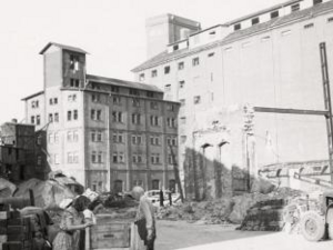 Ein schwarz-weiß Foto mit einem großen Gebäude. Vor dem Gebäude liegen Ruinen. Vorne links im Bild stehen zwei Frauen und ein Mann. 