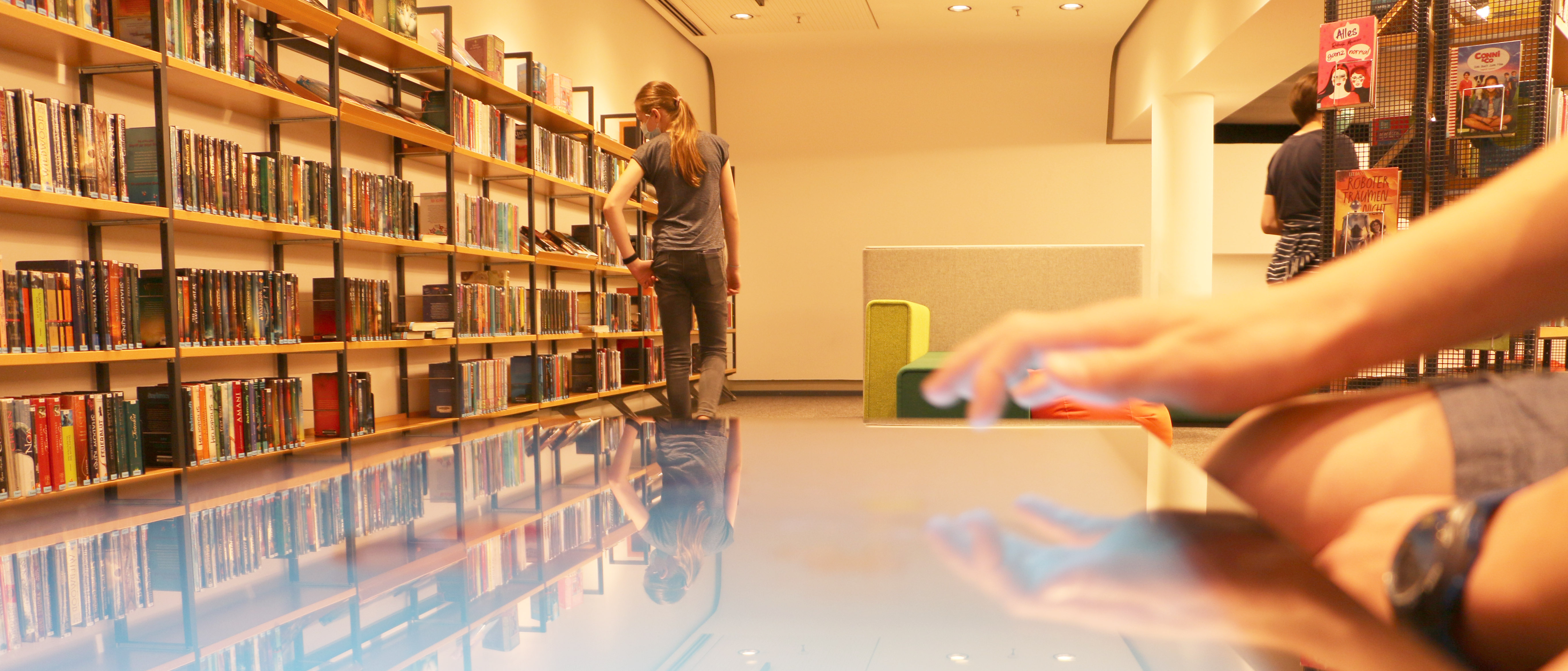 Im Vordergrund eine Hand an einem Touch-Screen; Im Hintergrund sucht ein Mädchen etwas in einem Bücherregal