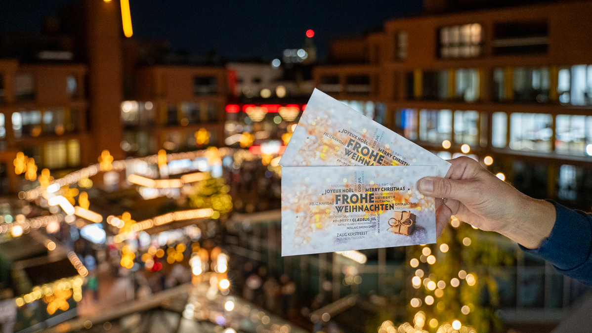 Blick auf den Weihnachtsmarkt im Ageidiimarkt mit zwei Gutscheinen der vhs Münster im Bildvordergrund.