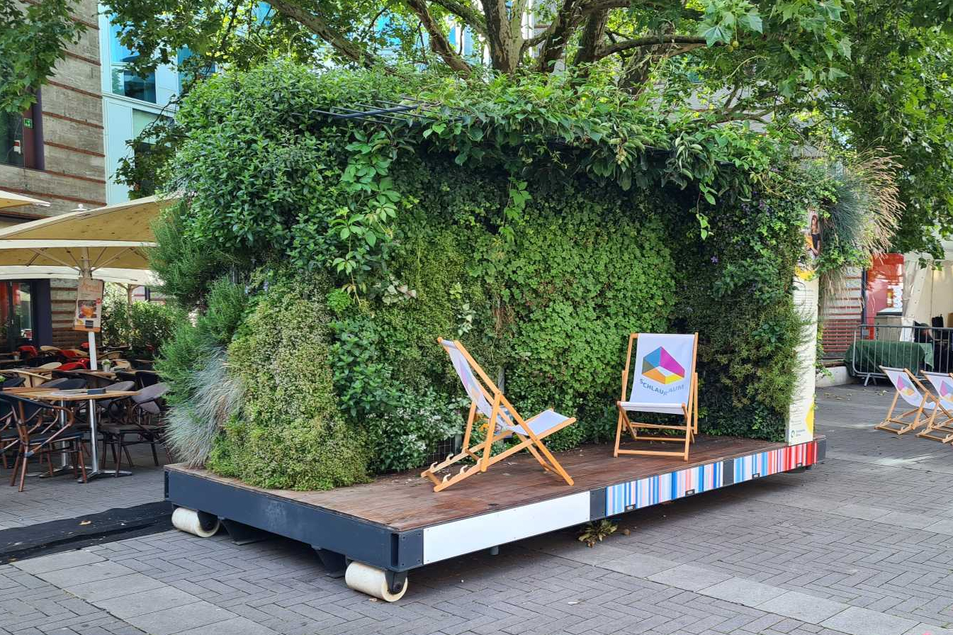 Bühne mit grüner Pflanzenwand und zwei Holzstühlen mit bunten Rückenlehnen auf gepflastertem Platz, im Hintergrund weitere Stuhlreihen und Bäume
