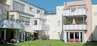 Foto der Hiltruper Wohngenossenschaft geschossen aus dem begrünten Innenhof heraus. Zu sehen ist das Gebäude mit heller Fassade, den Terassen und Balkonen.
