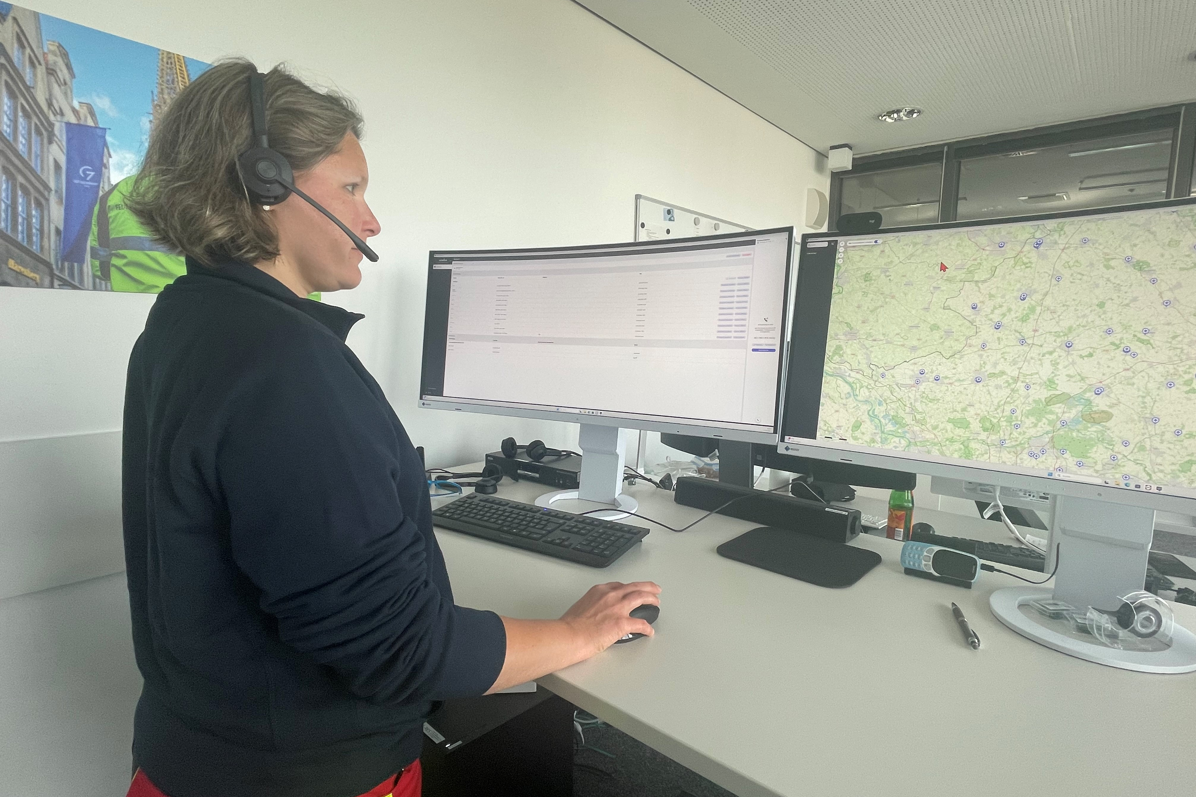 Eine Frau steht an einem Schreibtisch trägt ein Headset und guckt auf zwei Monitore.