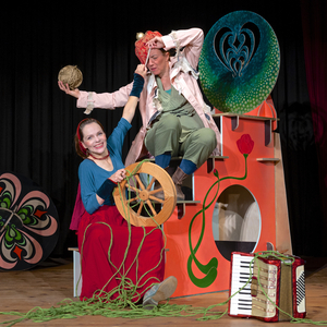 Eine Performerin sitzt auf einer Treppe im Bühnenbild und hält ein Wollknäul in der Hand. Die andere Performerin steht an einem Spinnrad.