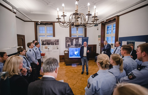 Ein Mann steht in der Mitte eines Ausstellungsraumes und redet. Um ihn herum stehen Polizist*innen in blauer Uniform. Sie hören zu. 