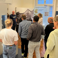 In einem Ausstellungsraum steht ein Mann neben einem großformatigen historischen Foto. Vor ihm stehen Menschen und hören zu.