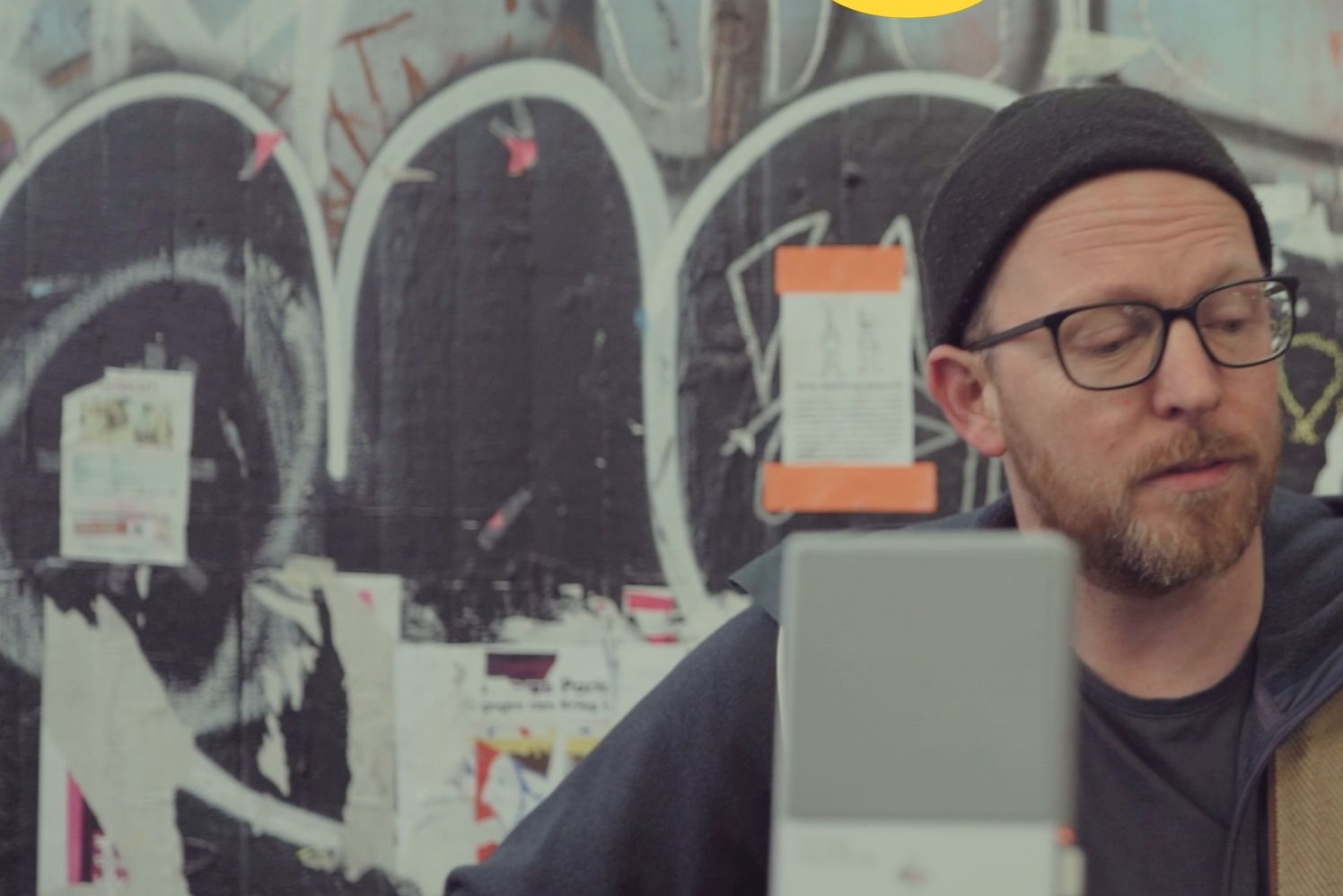 Mann mit Brille und Mütze spielt Gitarre vor einer mit Graffiti bedeckten Wand, darüber gelber Schriftzug 'honig'