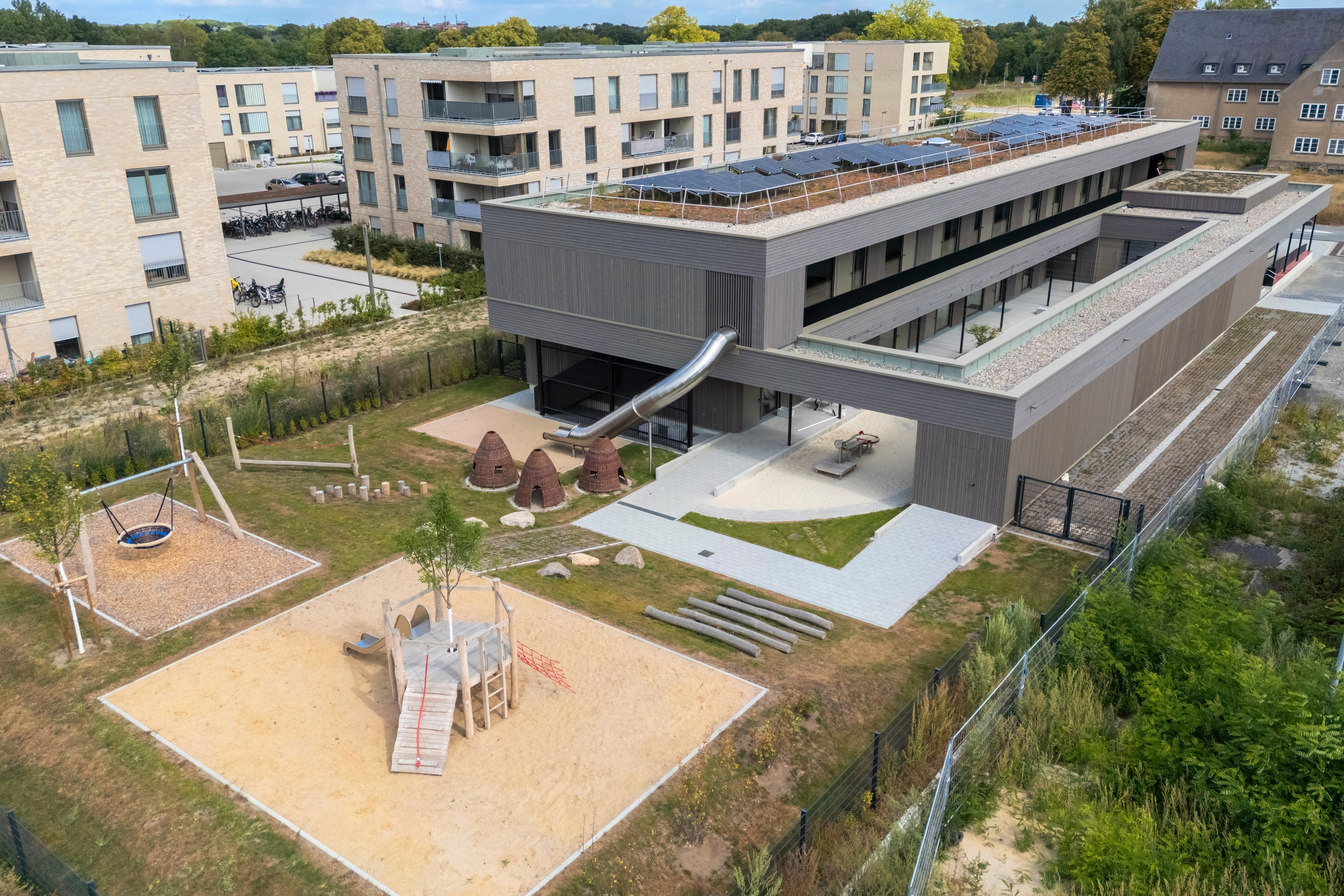 Blick von oben auf ein längliches Gebäude und einen mit Spielgeräten versehenen Außenbereich