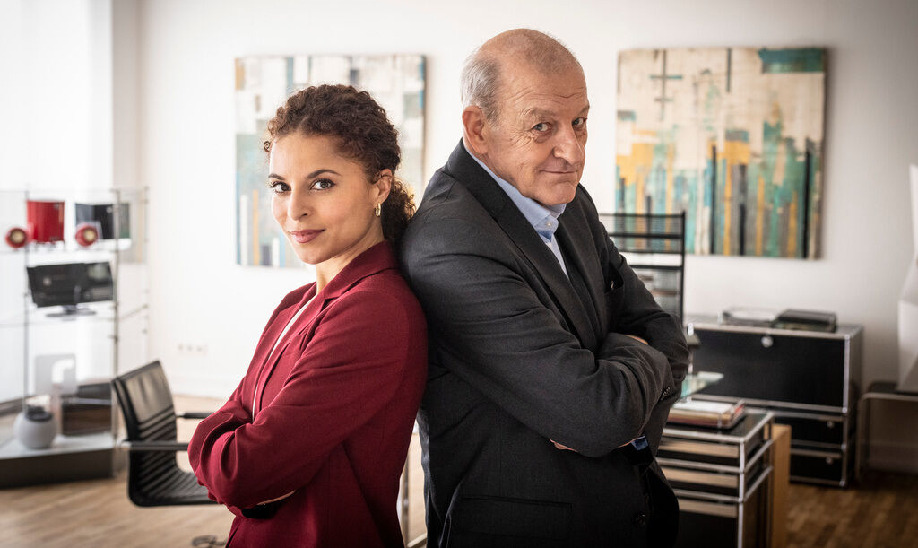 Patricia Meeden („Tessa Tilker“) und Leonard Lansink („Wilsberg“) stehen Rücken an Rücken in einem Büro und schauen in die Kamera.
