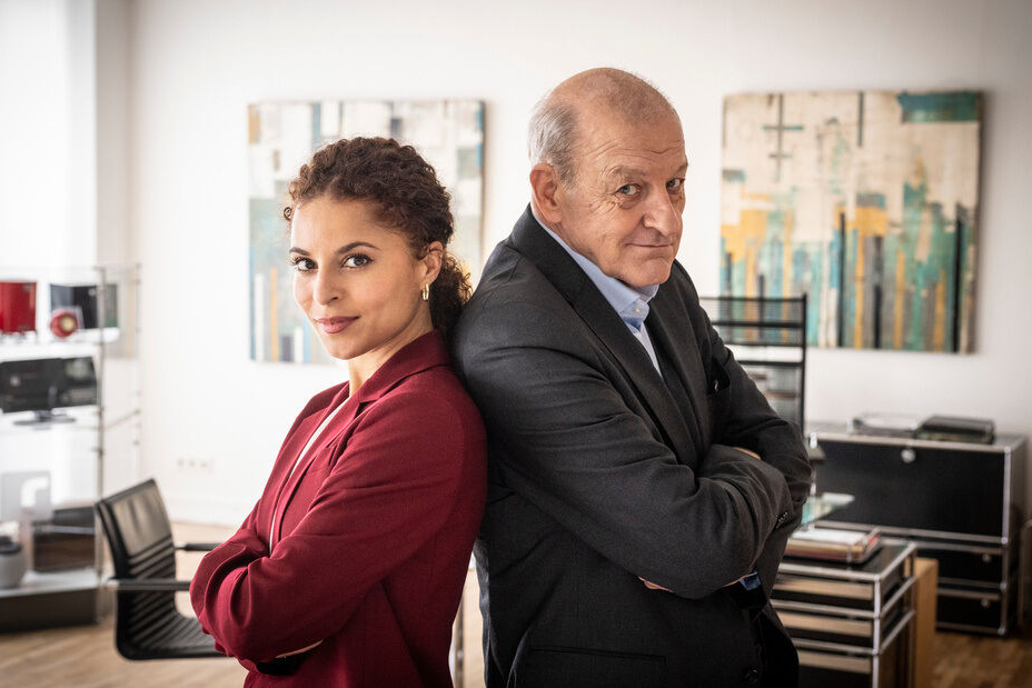 Patricia Meeden („Tessa Tilker“) und Leonard Lansink („Wilsberg“) stehen Rücken an Rücken in einem Büro und schauen in die Kamera.