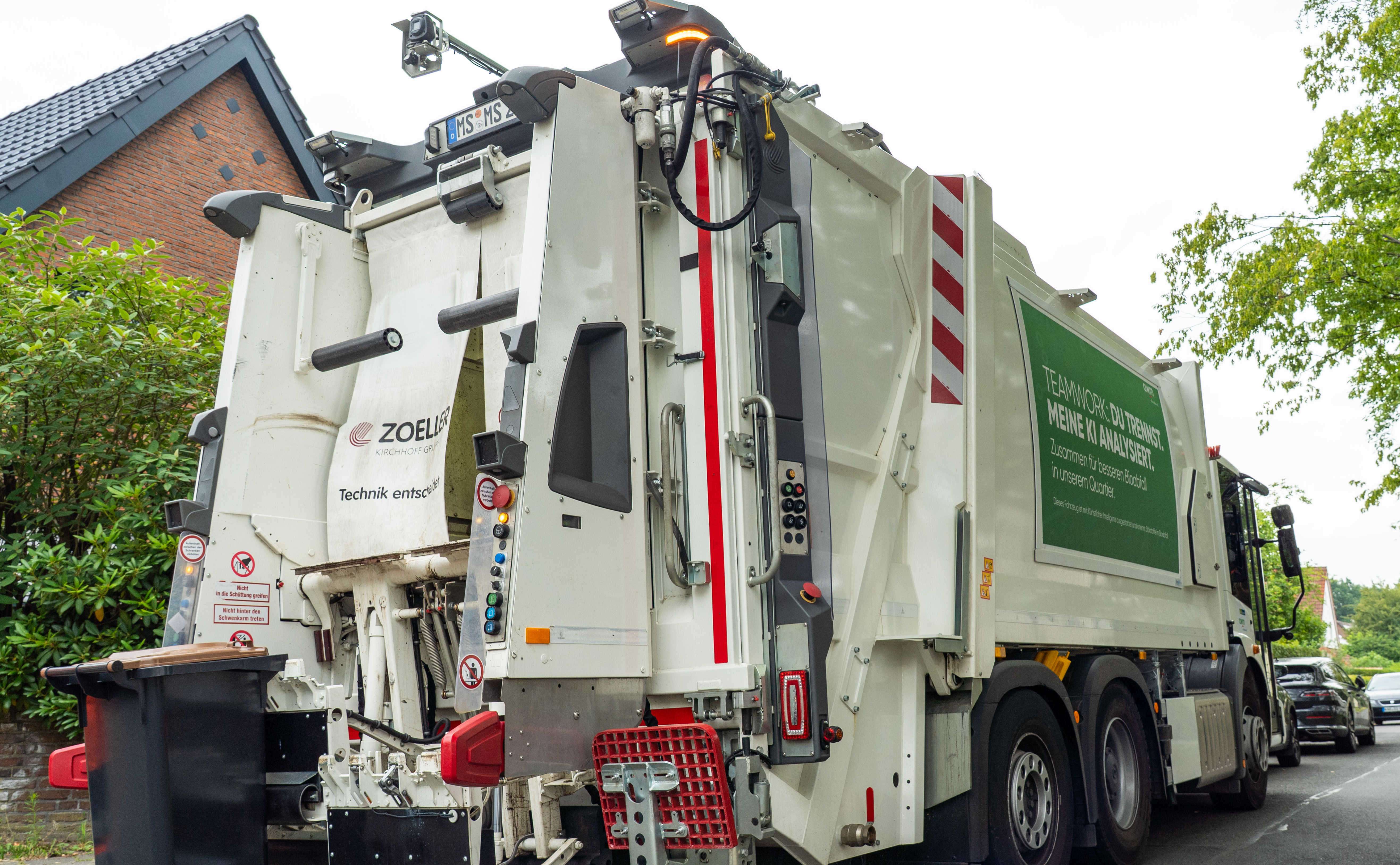 Ein großer heller Müllwagen vor einem Wohnhaus, der gerade eine Biotonne auflädt. Am Dach des des Müllwagens ist eine Kamera installiert, die die Entleerung der Tonne beobachten kann.