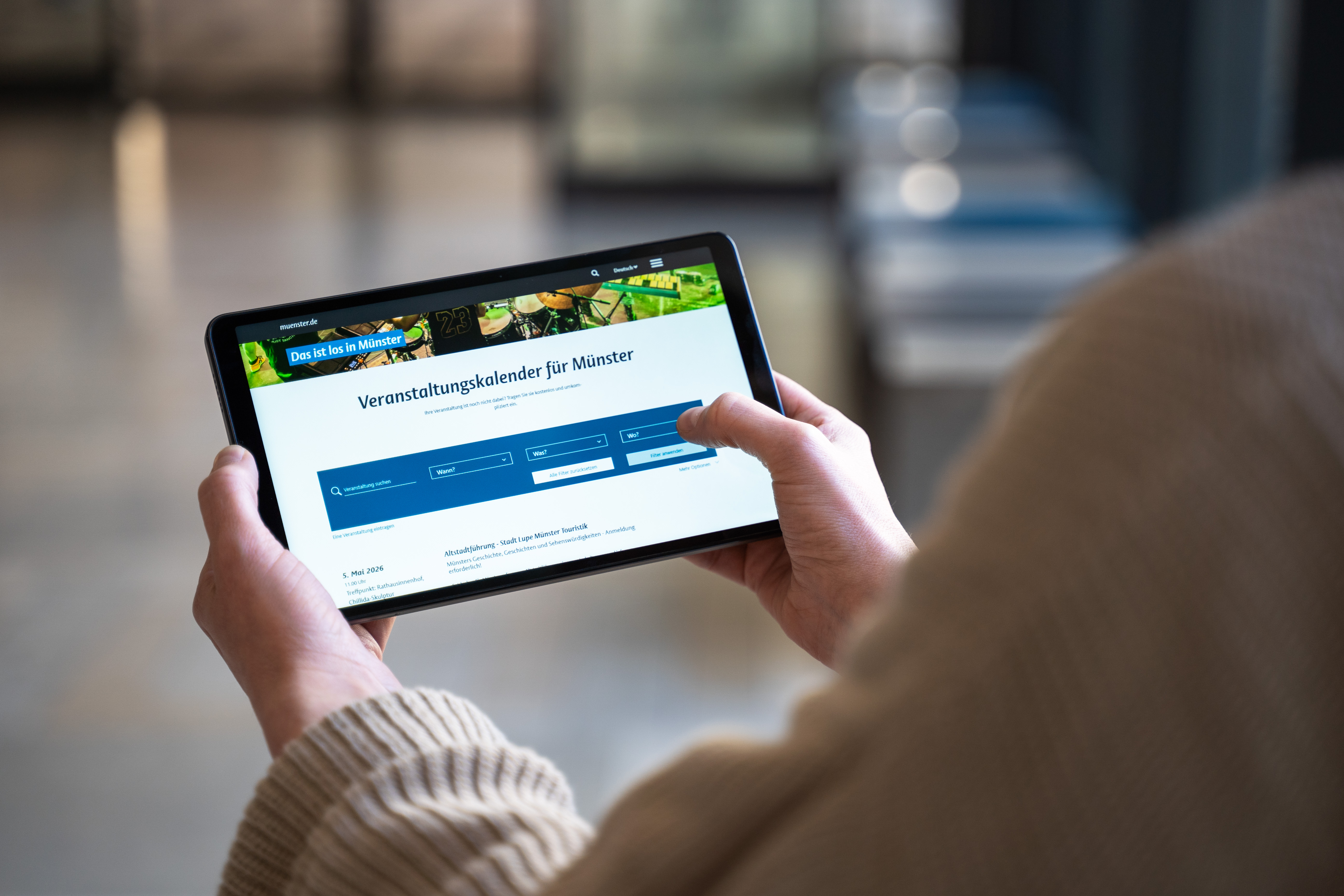 Eine Person, die nur von hinten zu sehen ist, hält ein Tablet auf dem der Veranstaltungskalender geöffnet ist. 