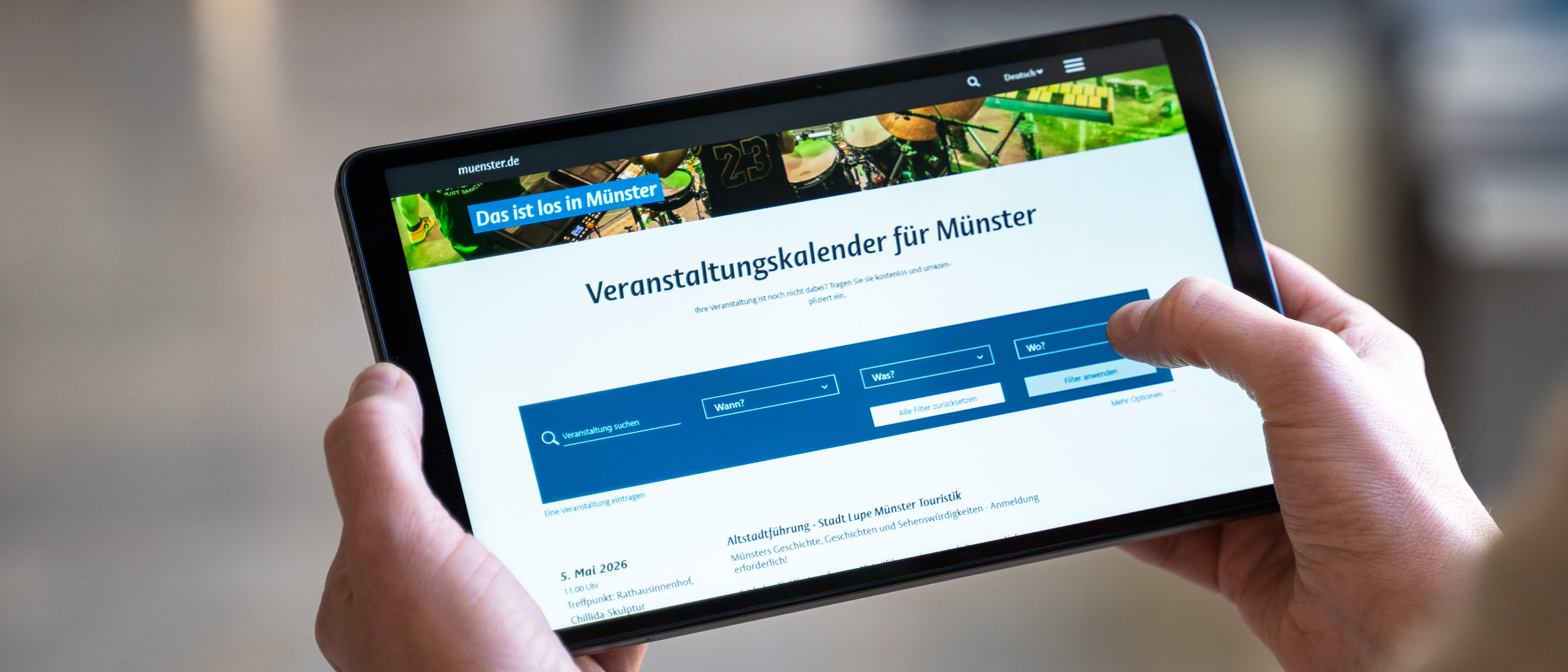 Eine Person, die nur von hinten zu sehen ist, hält ein Tablet auf dem der Veranstaltungskalender geöffnet ist. 