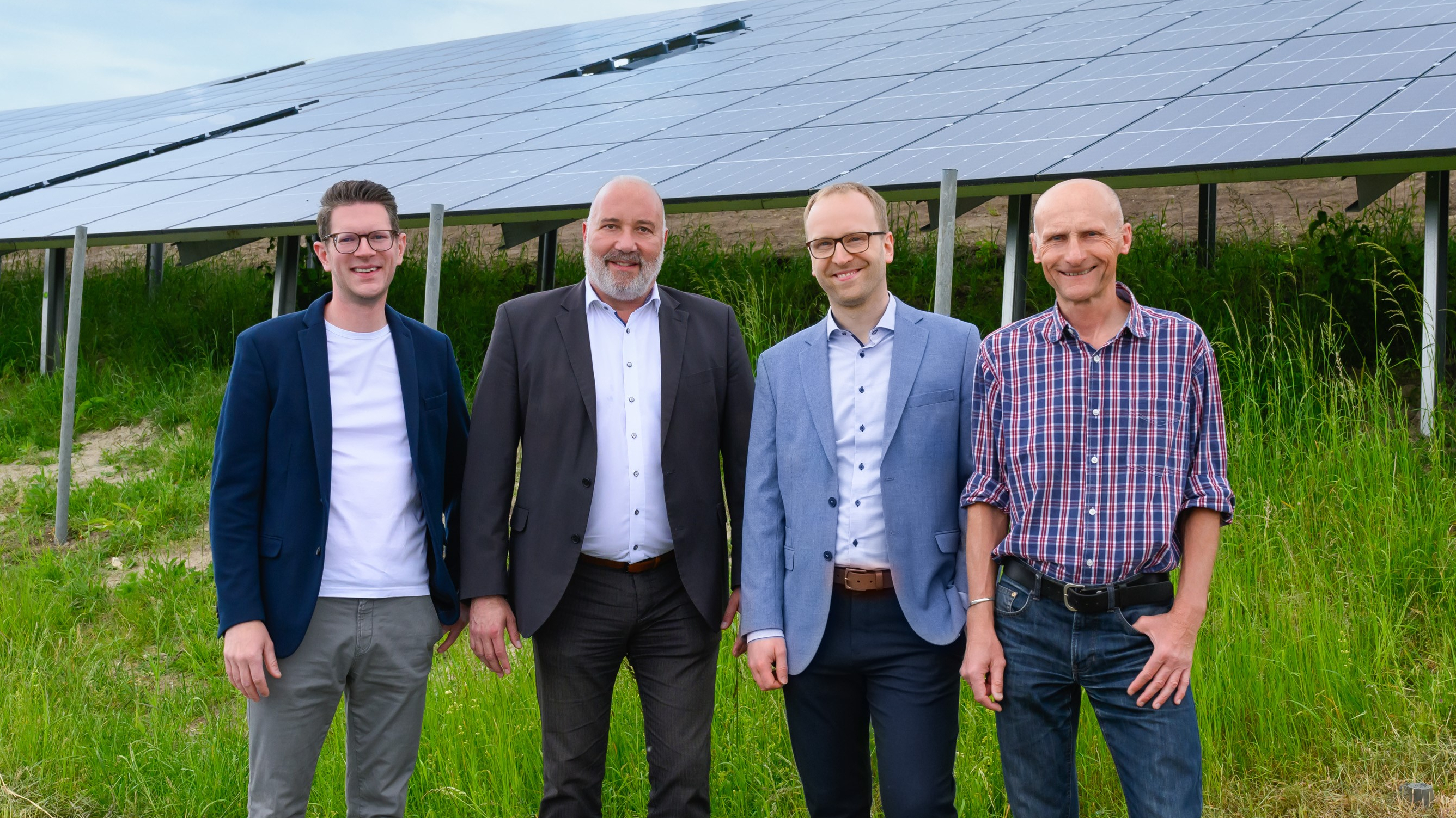 Die vier Herren stehen nebeneinander auf einem grünen Hügel, hinter ihnen sind viele Solarmodule.