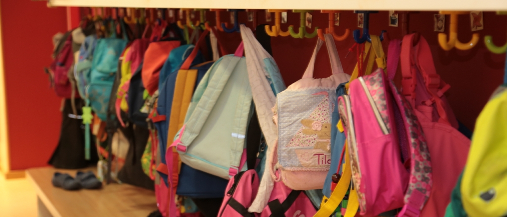 An einer Garderobe in einer Kita hängen Kinderrucksäcke und Kinderjacken in unterschiedlichen Farben.