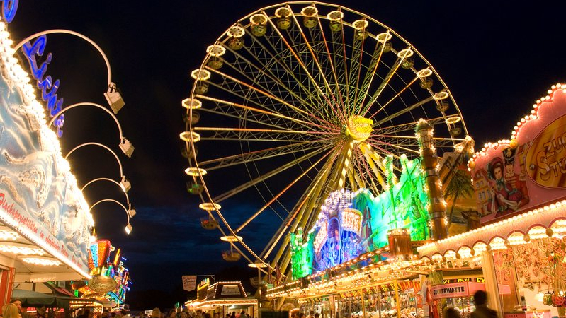 Eine Nachtausnahme vom Send in Münster. Am dunklen Himmel erscheint das beleuchtete, sich drehende Riesenrad sowie diverese illuminierte Verkaufsstände. Mehrere Besucher laufen zwischen den Ständen oder beobachten diverse Angebote.