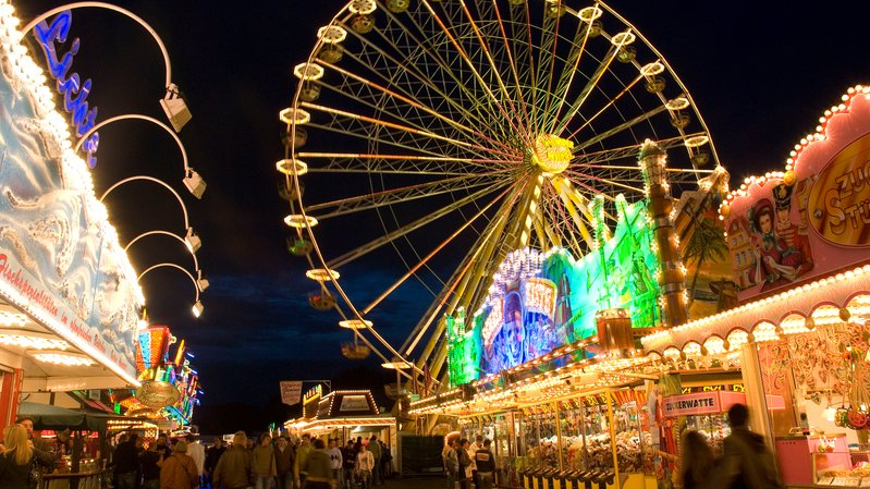 Eine Nachtausnahme vom Send in Münster. Am dunklen Himmel erscheint das beleuchtete, sich drehende Riesenrad sowie diverese illuminierte Verkaufsstände. Mehrere Besucher laufen zwischen den Ständen oder beobachten diverse Angebote.