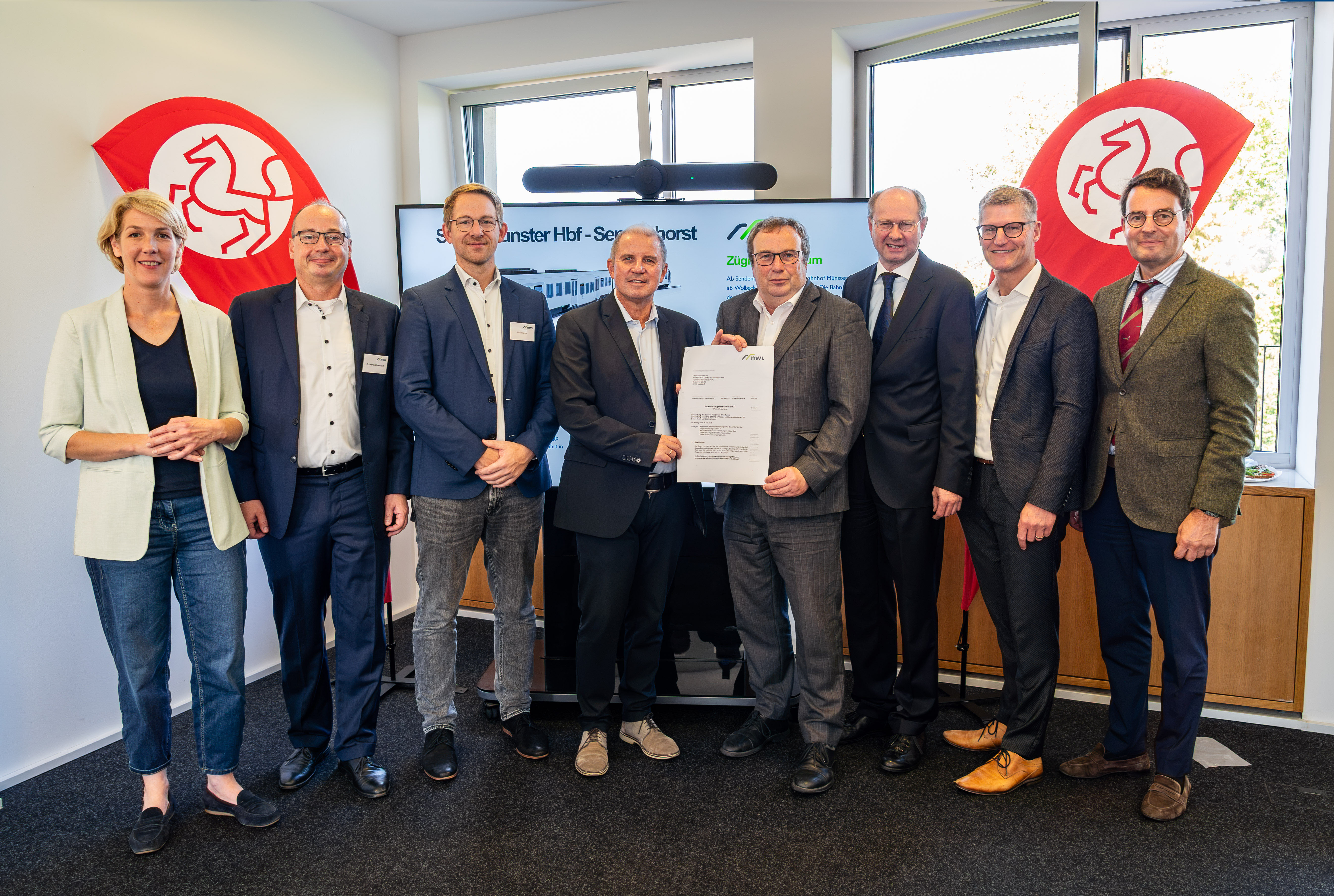 Katrin Reuscher (v.l.), Bürgermeisterin von Sendenhorst, Dr. Martin Uhlendorf, Leiter Infrastrukturförderung beim Zweckverband Nahverkehr Westfalen-Lippe (NWL), Nils Werner, Leiter Geschäftsbereich Markt & Kunde beim Zweckverband Nahverkehr Westfalen-Lippe (NWL), WLE-Geschäftsführer Detlef Berndt, NRW-Verkehrsminister Oliver Krischer, Landrat Dr. Olaf Gericke, Münsters Oberbürgermeister Tilman Fuchs und Regierungspräsident Andreas Bothe bei der Übergabe des Förderbescheids. Sie stehen in einem Bürorraum, im Hintergrund sind zwei Fahnen mit Pferde-Icon zu sehen. 
