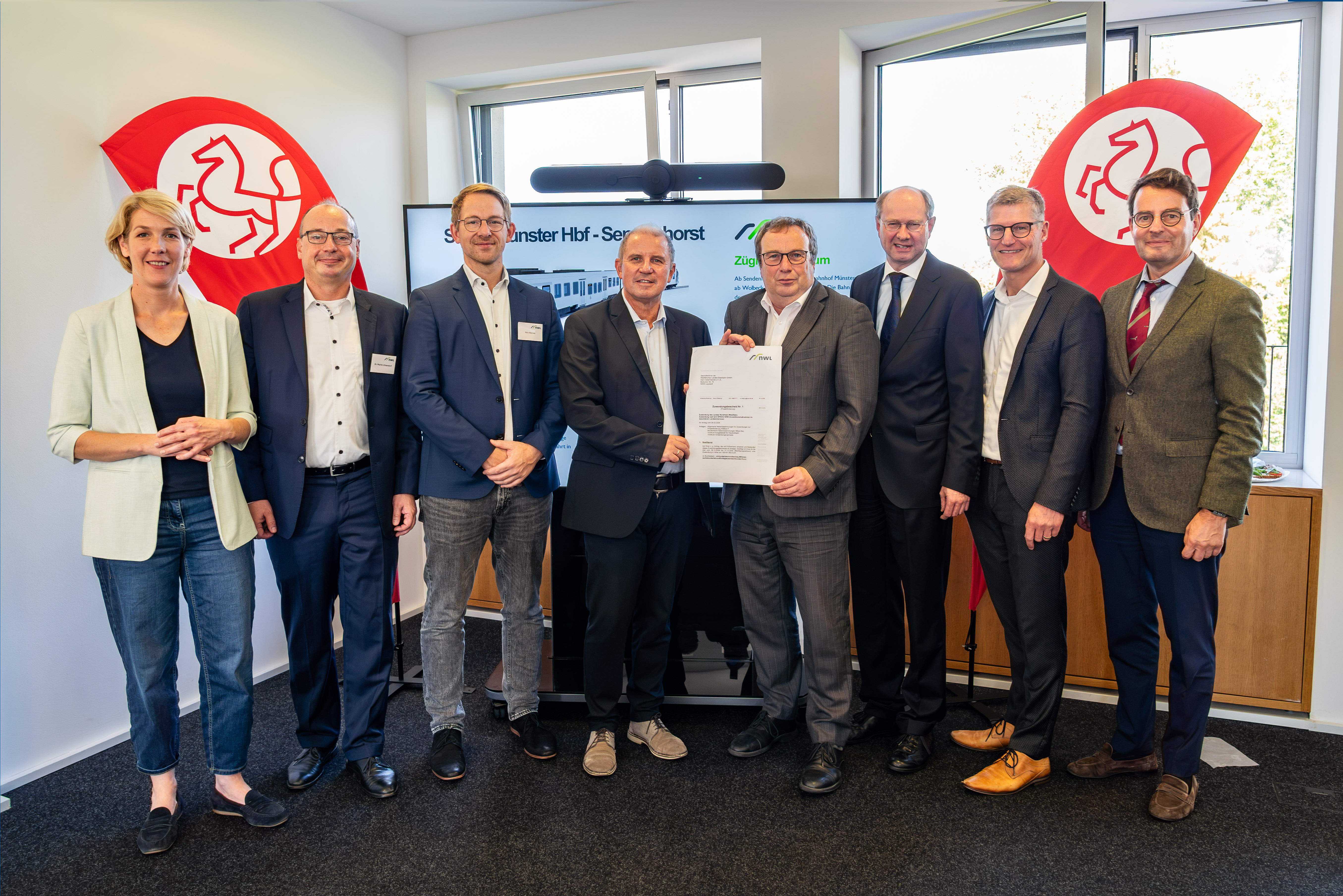 Katrin Reuscher (v.l.), Bürgermeisterin von Sendenhorst, Dr. Martin Uhlendorf, Leiter Infrastrukturförderung beim Zweckverband Nahverkehr Westfalen-Lippe (NWL), Nils Werner, Leiter Geschäftsbereich Markt & Kunde beim Zweckverband Nahverkehr Westfalen-Lippe (NWL), WLE-Geschäftsführer Detlef Berndt, NRW-Verkehrsminister Oliver Krischer, Landrat Dr. Olaf Gericke, Münsters Oberbürgermeister Tilman Fuchs und Regierungspräsident Andreas Bothe bei der Übergabe des Förderbescheids. Sie stehen in einem Bürorraum, im Hintergrund sind zwei Fahnen mit Pferde-Icon zu sehen. 