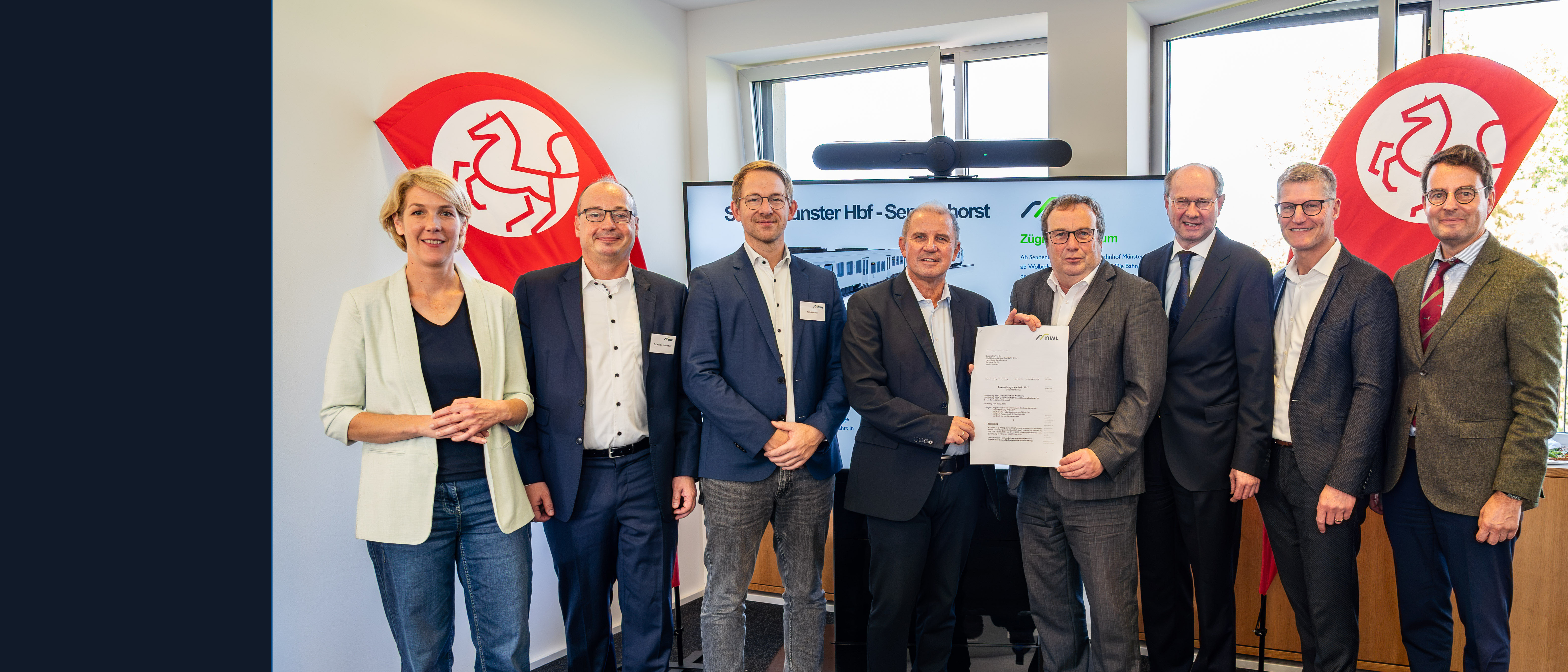 Katrin Reuscher (v.l.), Bürgermeisterin von Sendenhorst, Dr. Martin Uhlendorf, Leiter Infrastrukturförderung beim Zweckverband Nahverkehr Westfalen-Lippe (NWL), Nils Werner, Leiter Geschäftsbereich Markt & Kunde beim Zweckverband Nahverkehr Westfalen-Lippe (NWL), WLE-Geschäftsführer Detlef Berndt, NRW-Verkehrsminister Oliver Krischer, Landrat Dr. Olaf Gericke, Münsters Oberbürgermeister Tilman Fuchs und Regierungspräsident Andreas Bothe bei der Übergabe des Förderbescheids. Sie stehen in einem Bürorraum, im Hintergrund sind zwei Fahnen mit Pferde-Icon zu sehen. 