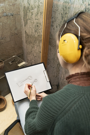 Eine Frau trägt einen gelben Gehörschutz. Sie sitzt auf einem Hocker vor einer bodentiefen Vitrine im Ausstellungsraum. Auf ihrem Schoß liegt ein Klemmbrett auf dem sie ein Ausstellungsobjekt nachzeichnet.