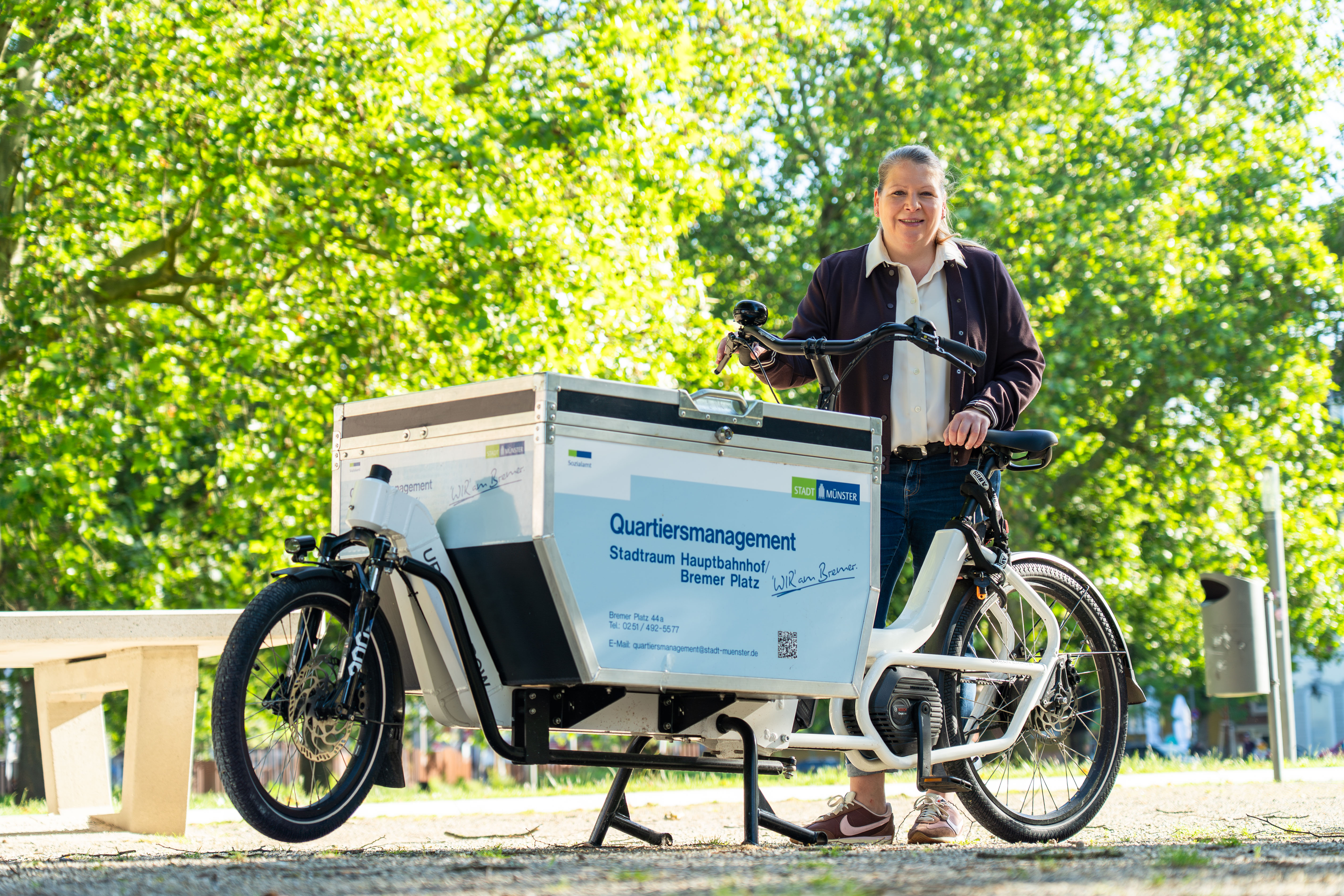 Judith Döpker steht in der Parkanlage am Bremer Platz hinter einem Lastenrad, begrünte Bäume im Hintergrund.