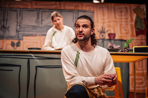 Der Performer sitzt in der Hocke im Vordergrund. Die Performerin steht hinter ihm hinter einer Küchenanrichte und schaut ihn an.