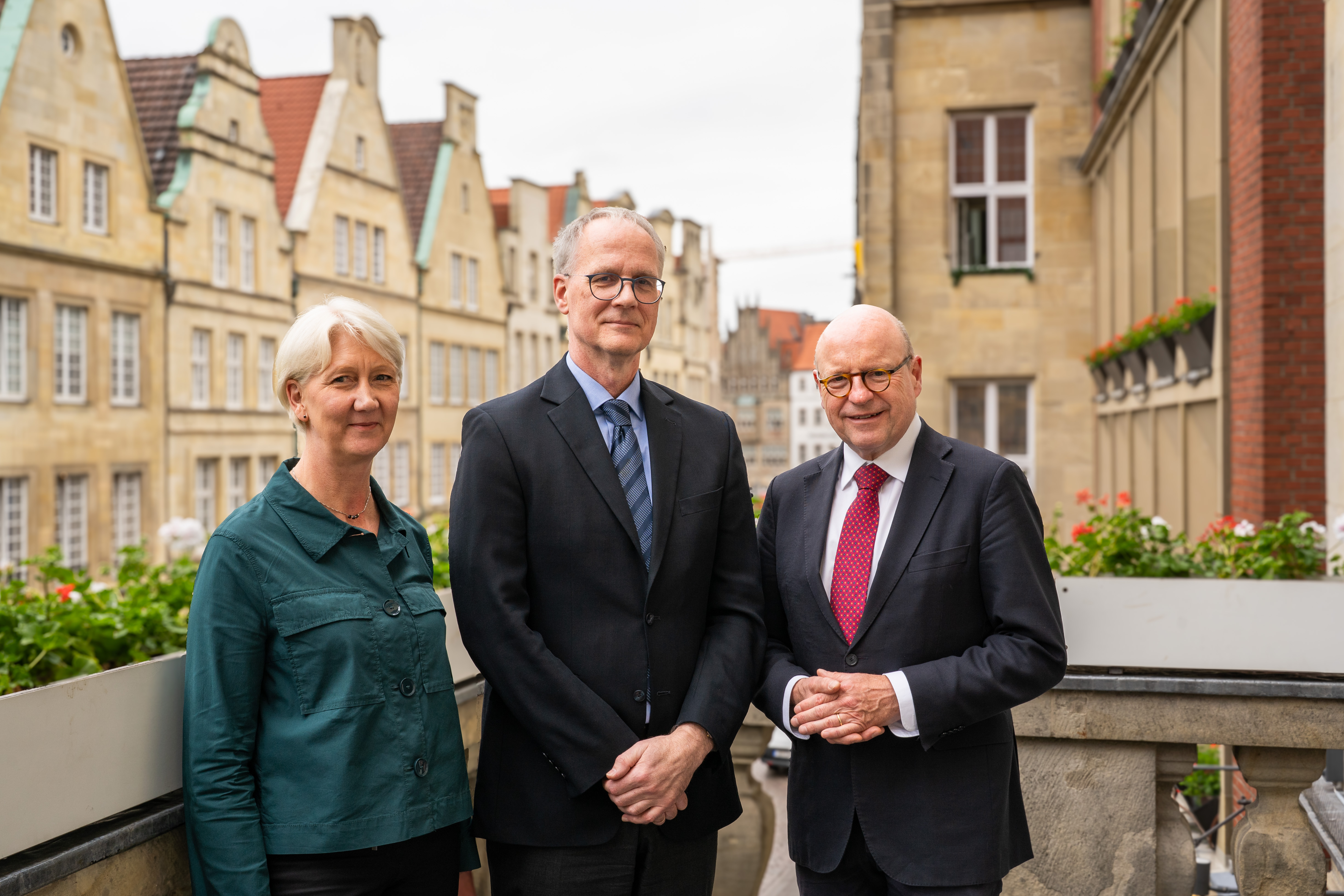 Die drei Personen stehen auf dem Balkon des historischen Rathauses in Münster, im Hintergrund sind die kunstvollen Außenfassaden des Prinzipalmarktes zu sehen.